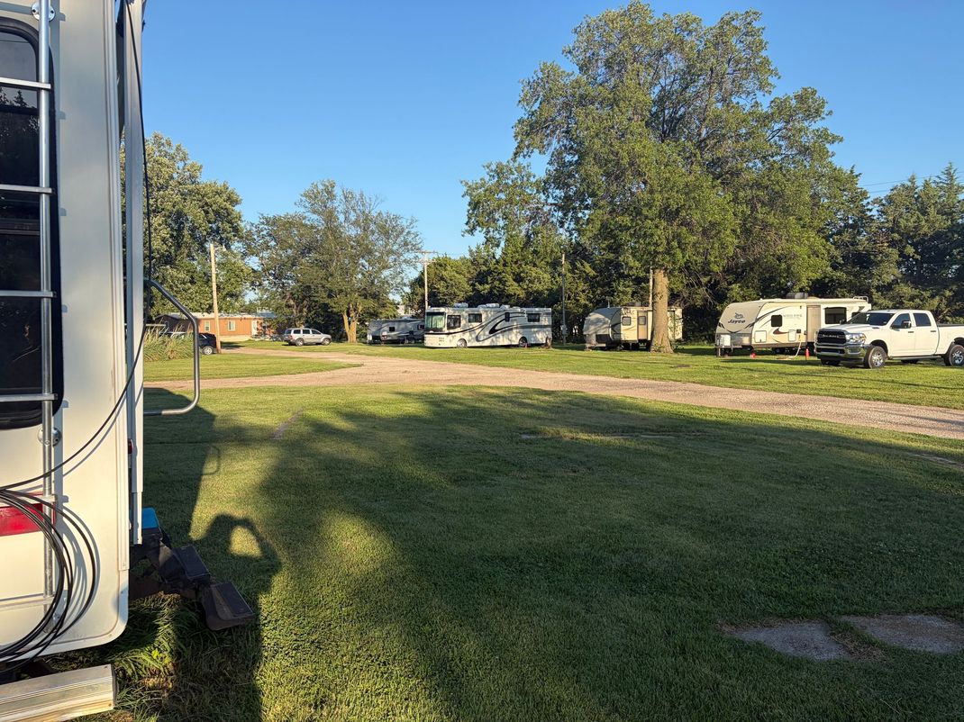 Grassy RV park with parked trailers, trees, and a dirt path on a sunny day