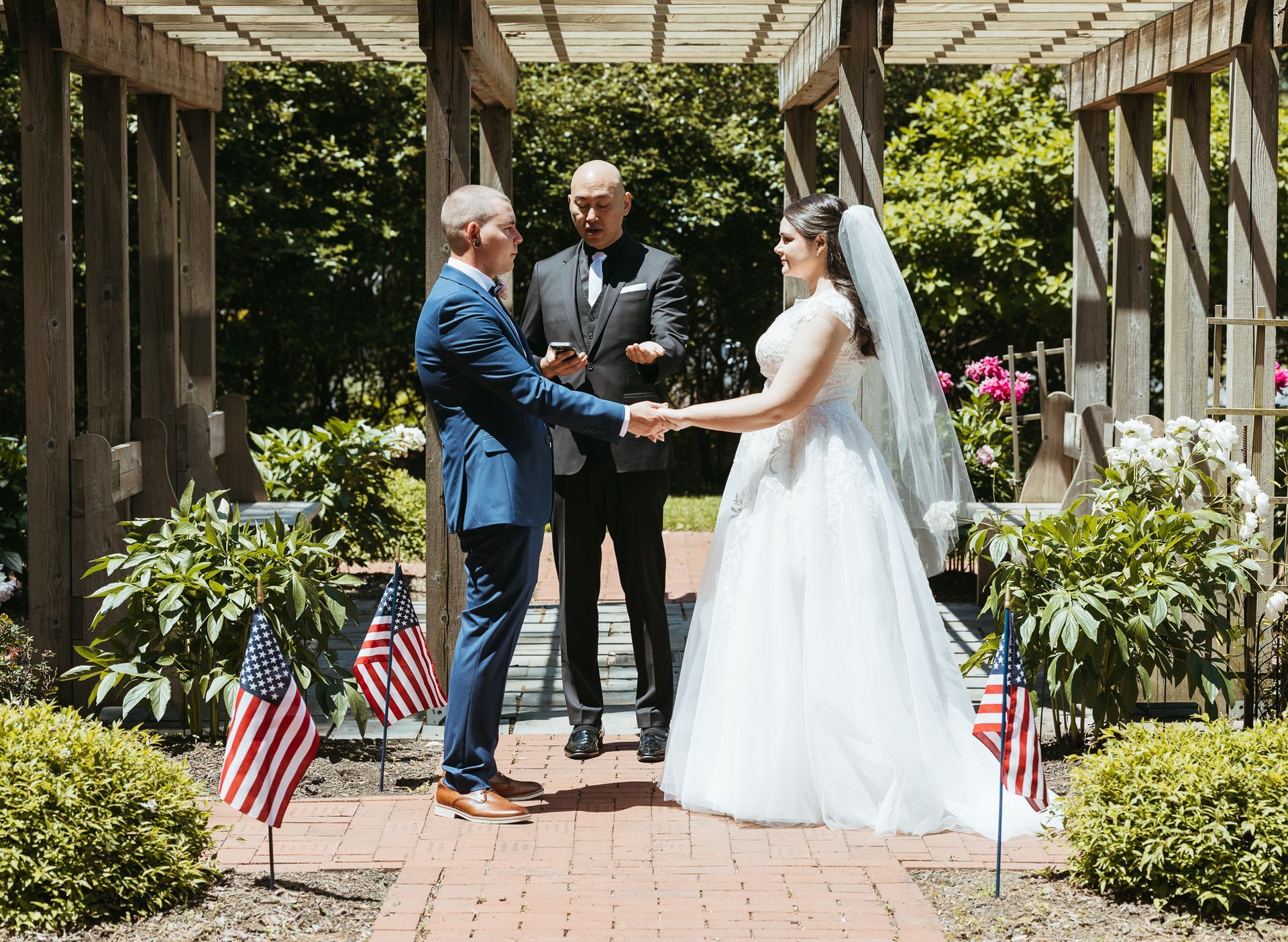 An officiant performs an outdoor wedding ceremony for a couple holding hands beneath a wooden pergola with U.S. flags.