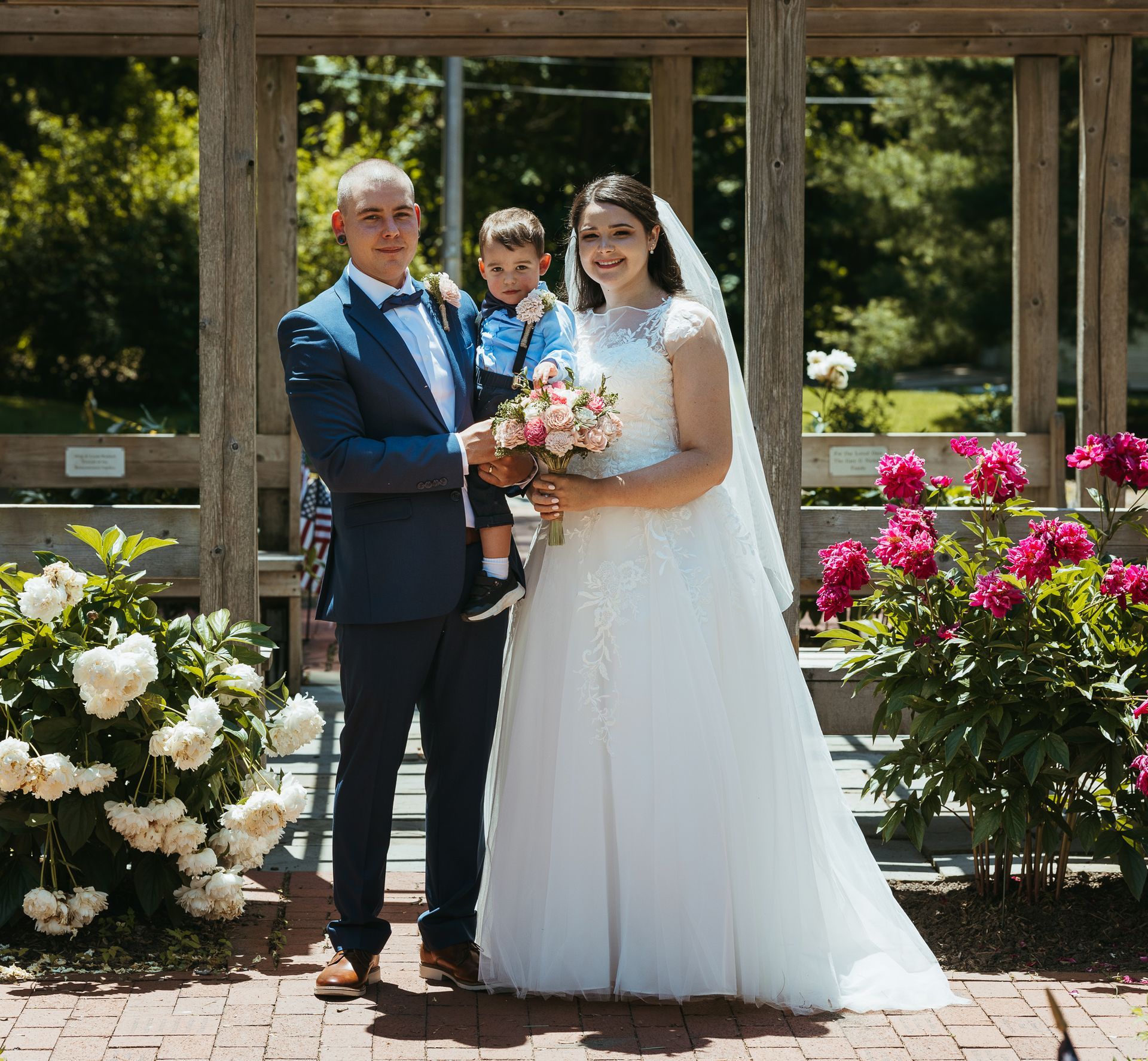 A bride and groom holding a young child, standing in a garden with white and pink flowers in front of a wooden pergola.