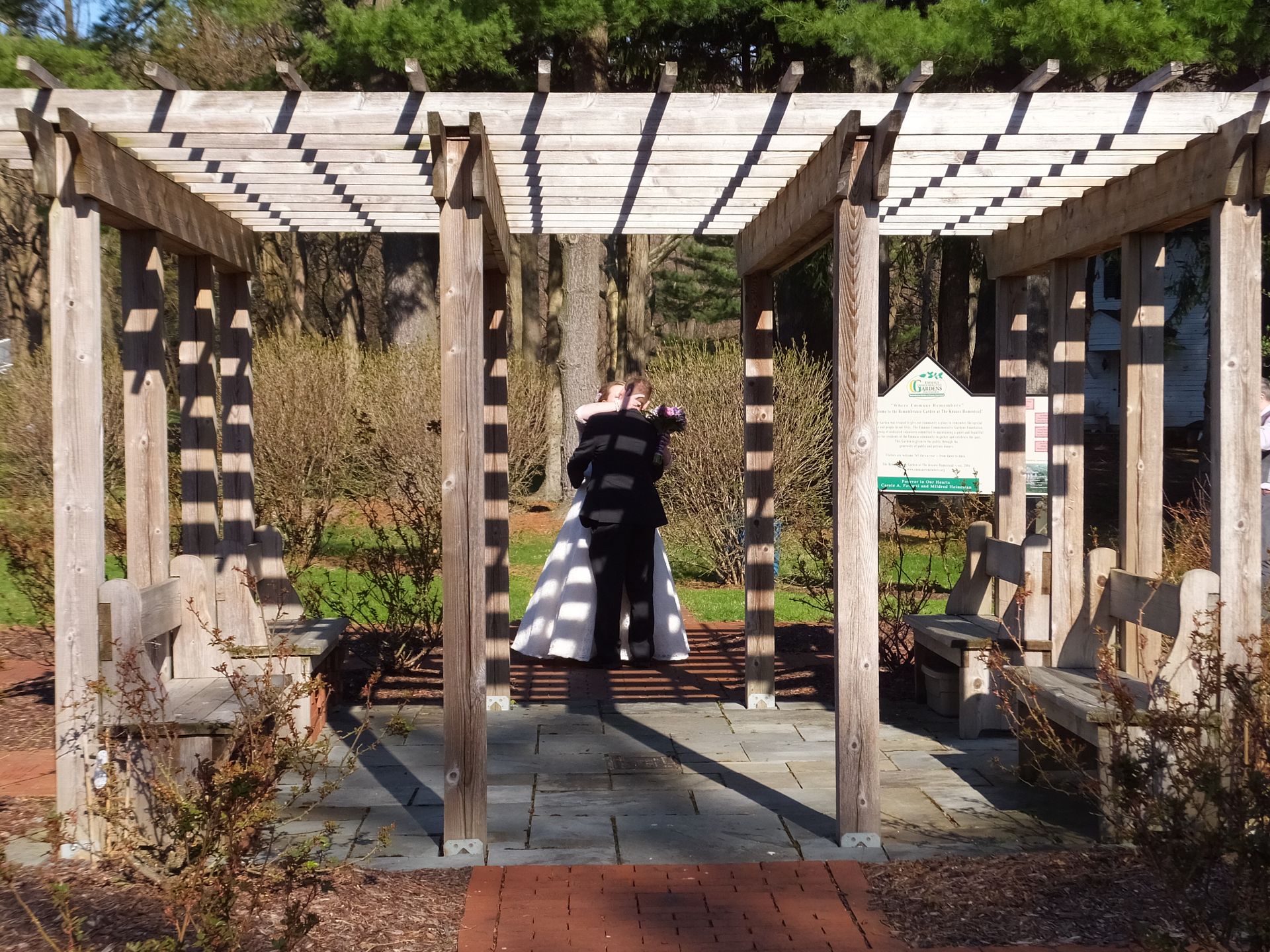A couple embracing under a wooden pergola on a stone patio in a park.