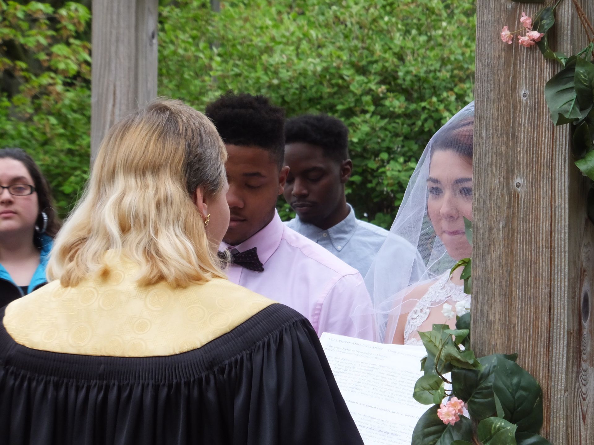 A wedding ceremony officiant stands before a couple in formal wear, framed by foliage in an outdoor setting.