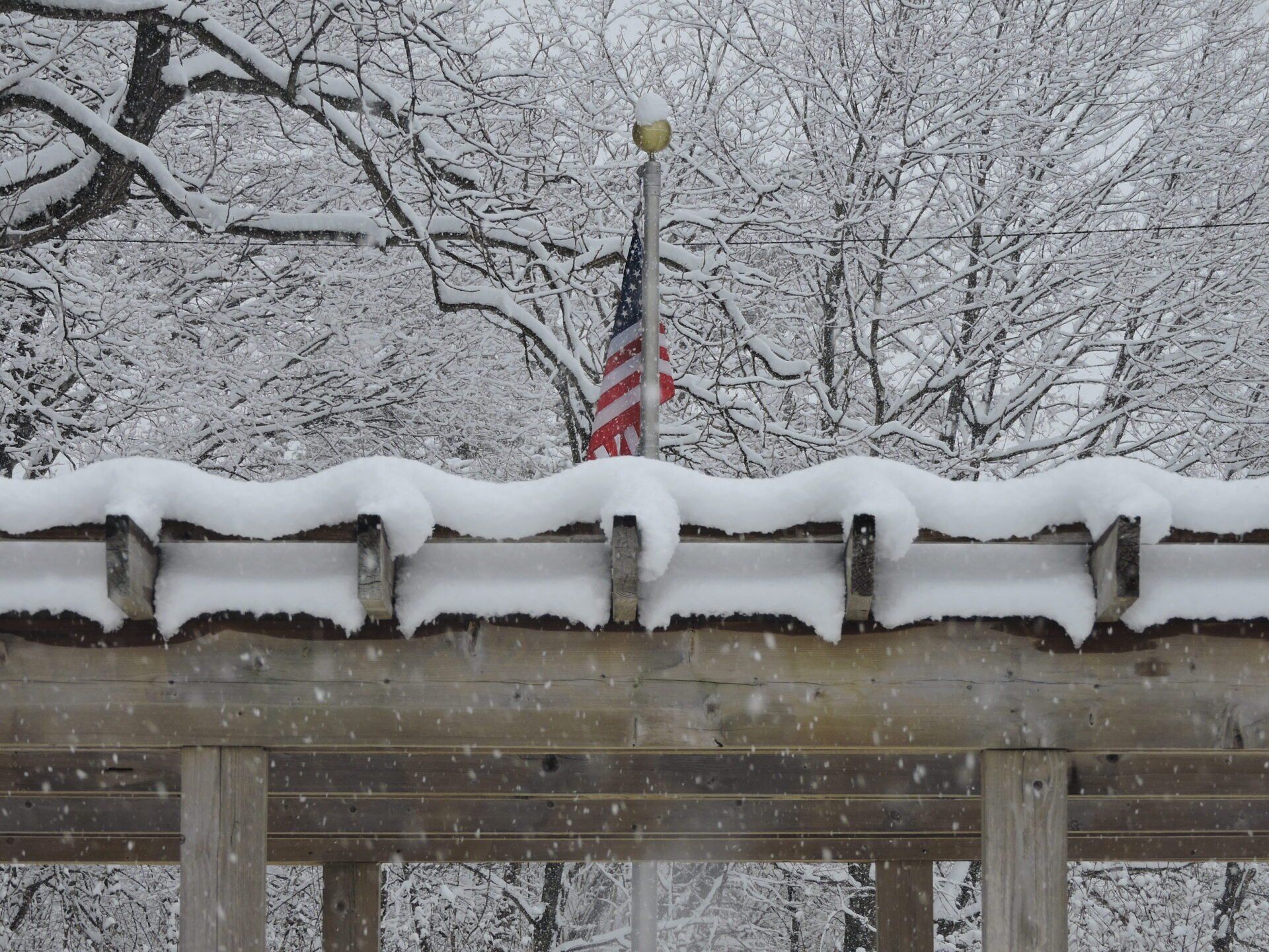 A wooden pergola structure topped with thick, fresh snow, with an American flag visible in the background trees.