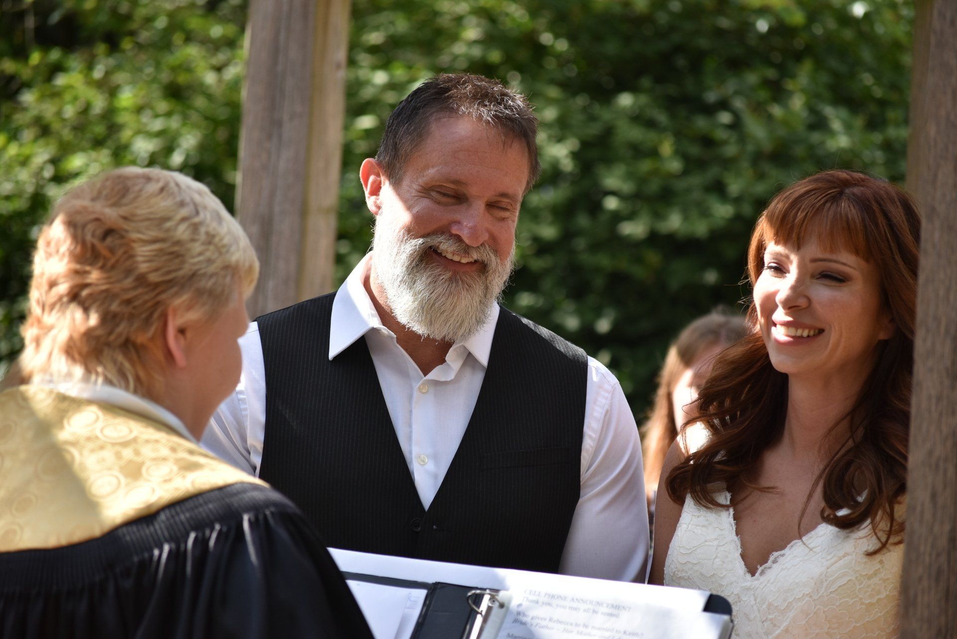 A couple smiles during an outdoor wedding ceremony, standing before an officiant in a black robe.