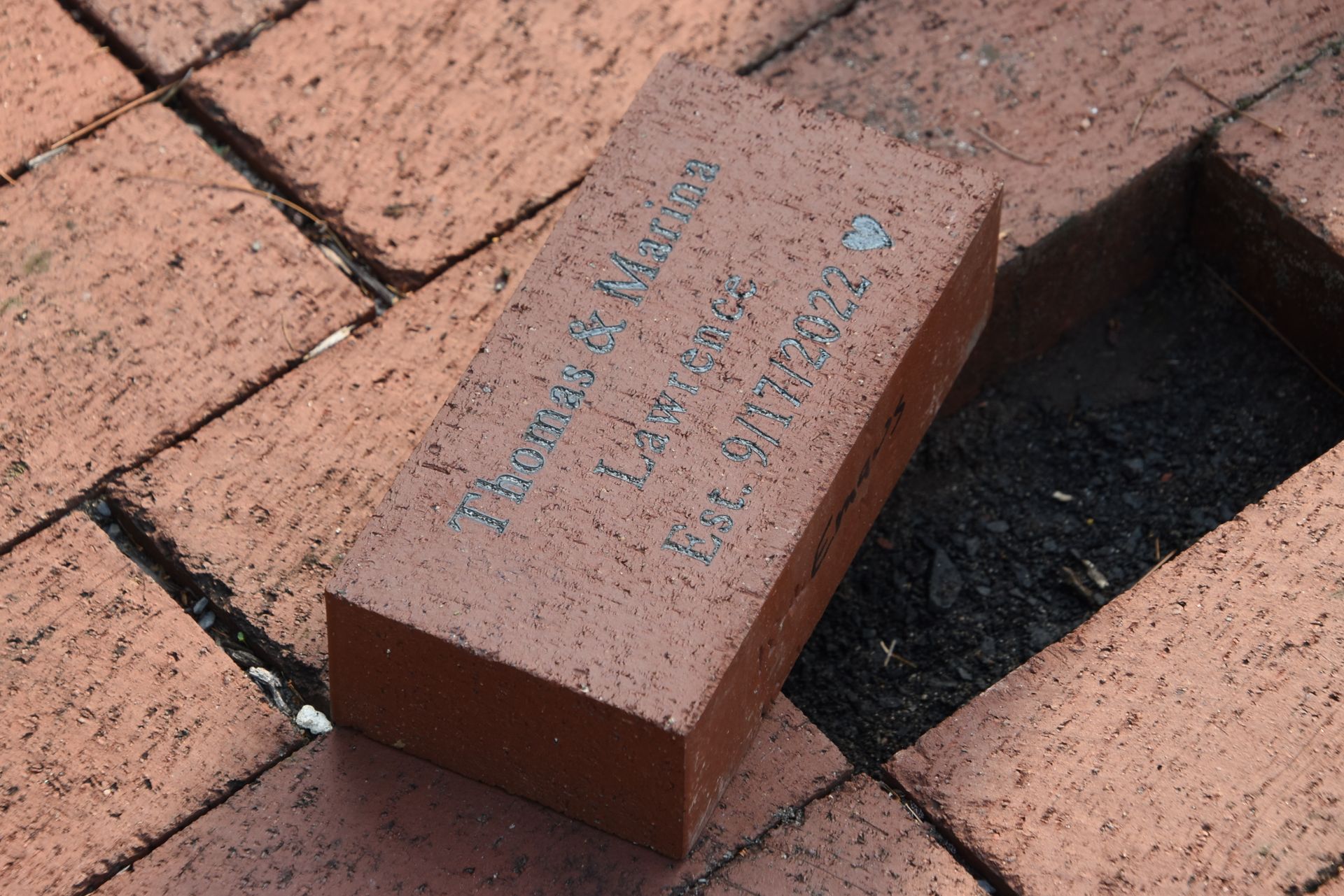 A red brick with an engraved name and date, sitting partially out of a hole in a paved brick walkway.