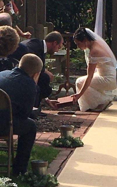 A couple kneeling on a brick path at an outdoor wedding ceremony, placing an object into a wooden box.