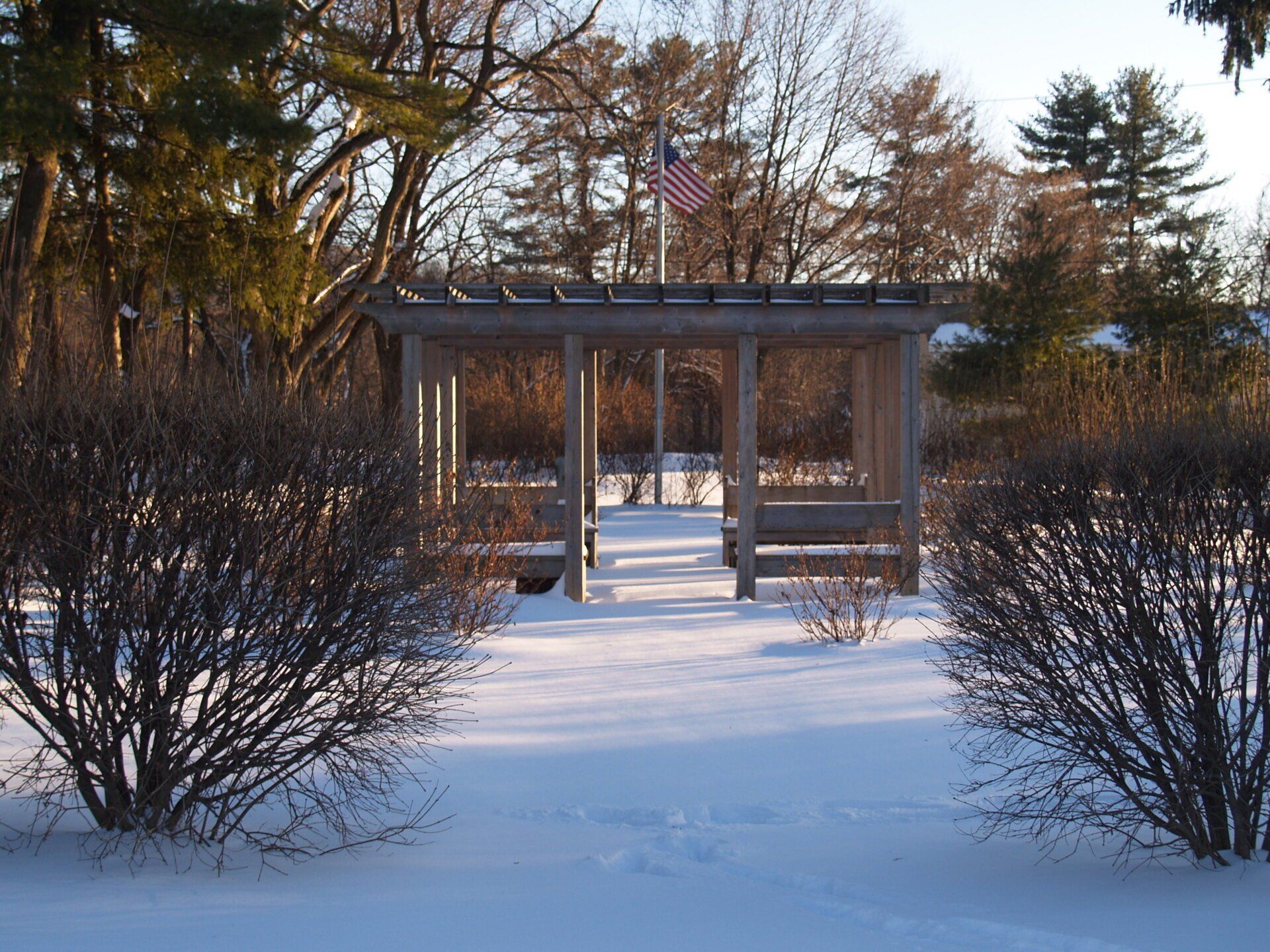 A wooden pergola stands in a snow-covered park, with an American flag flying on a pole behind it.