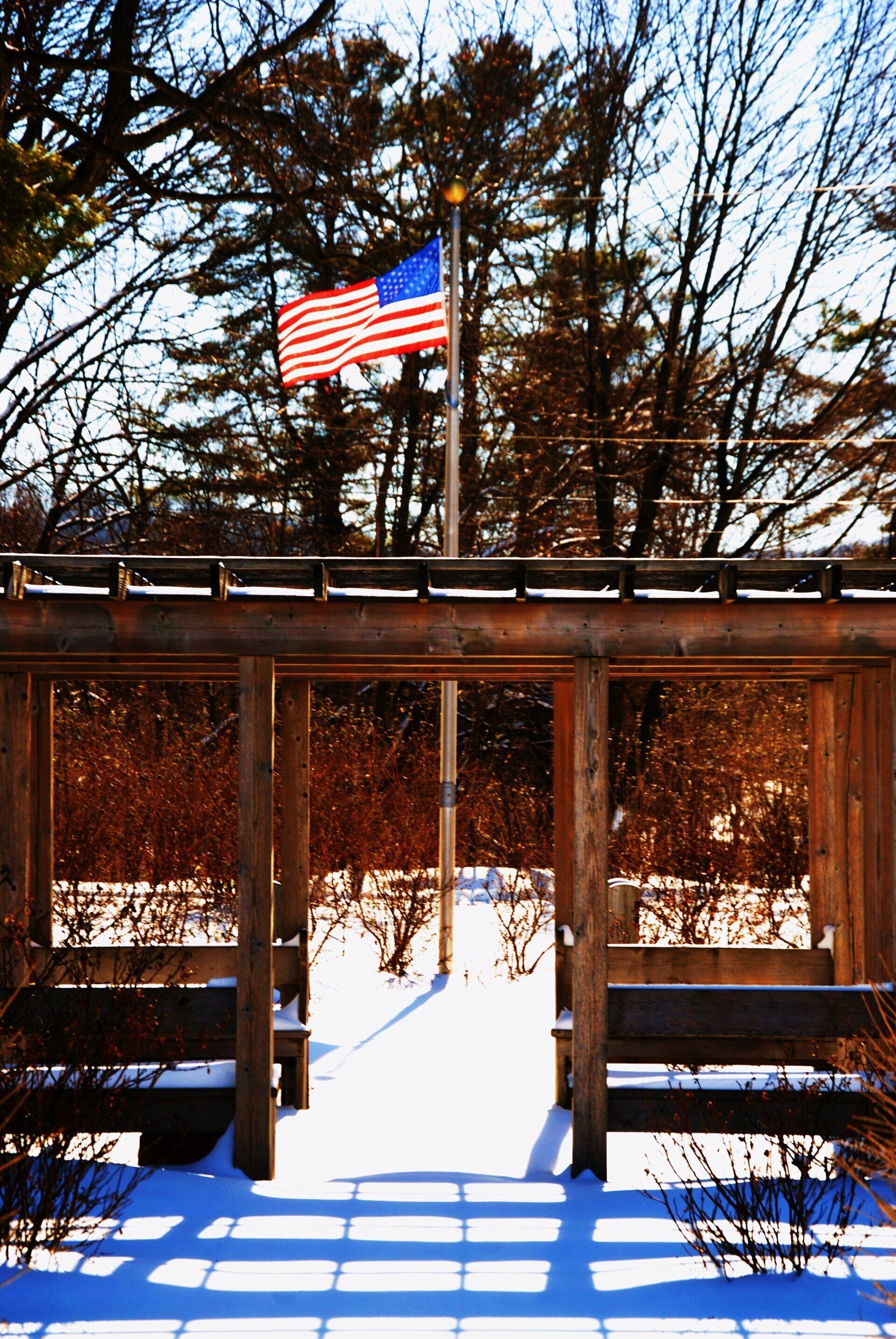 American flag flying above a wooden pergola covered in snow, surrounded by trees on a sunny winter day.