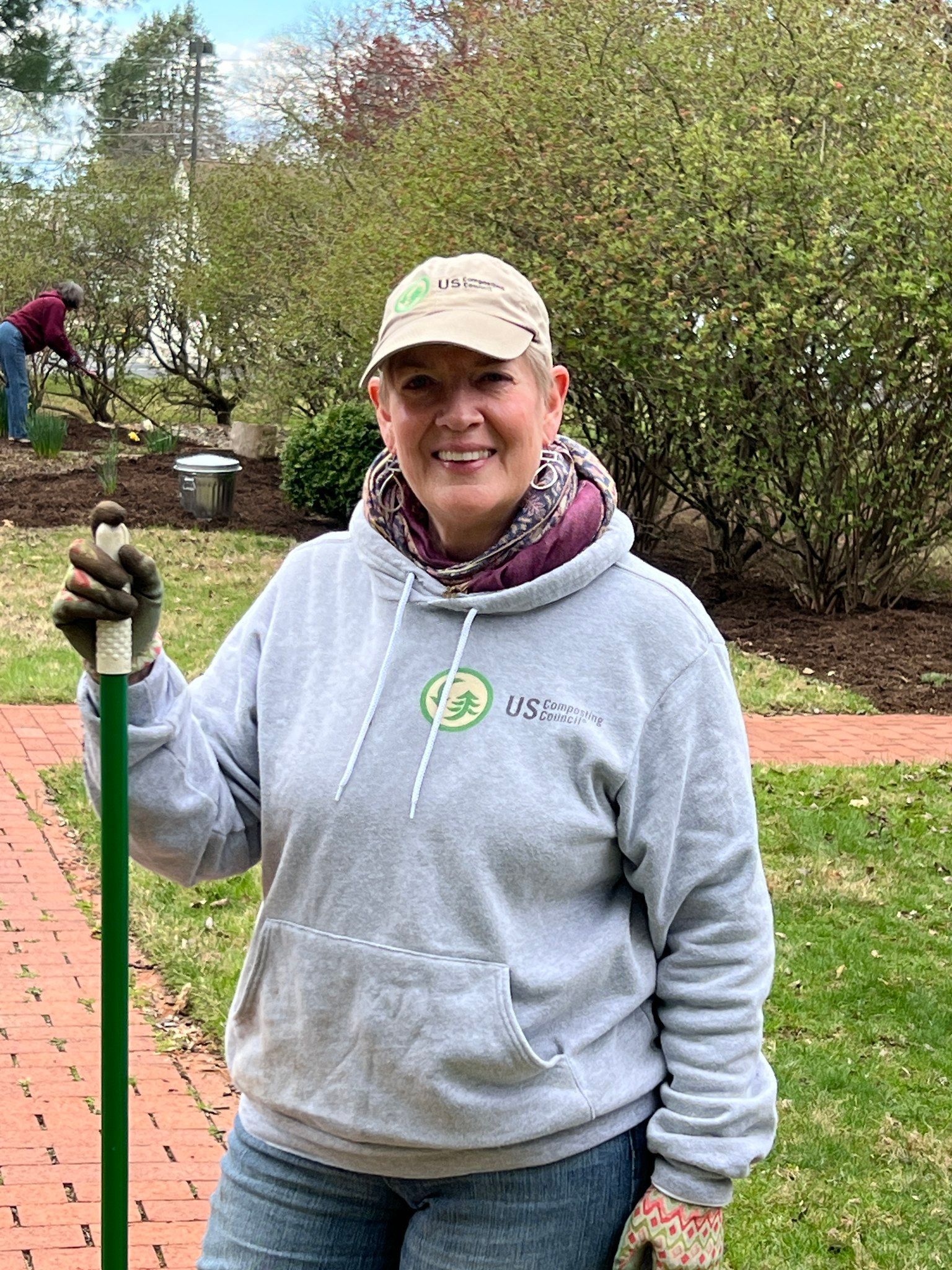 A smiling person in a gray hooded sweatshirt and tan cap stands outdoors holding a garden tool, with trees in the background.