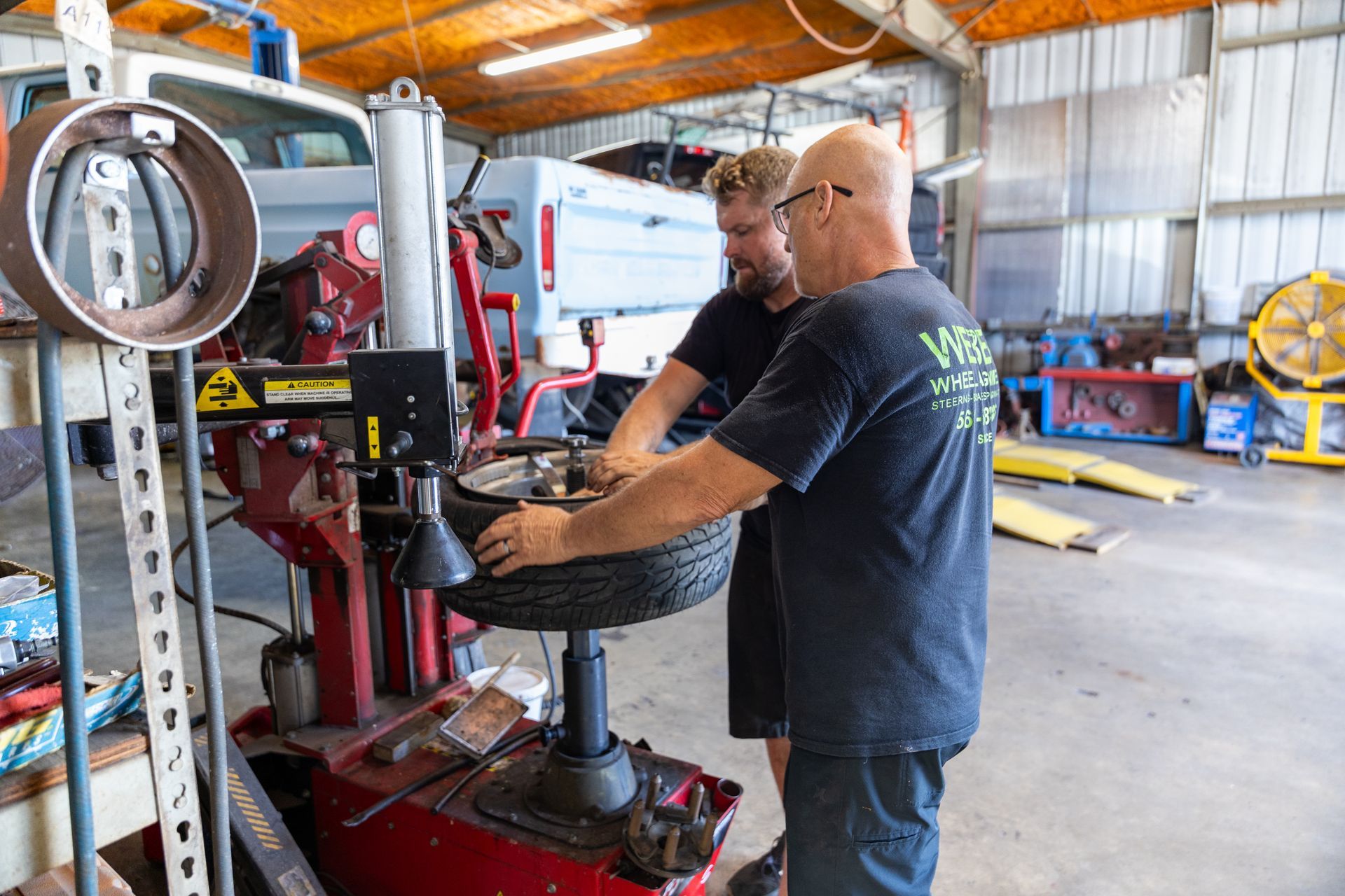 Two mechanics working on a tire machine in a garage with tools and equipment around them