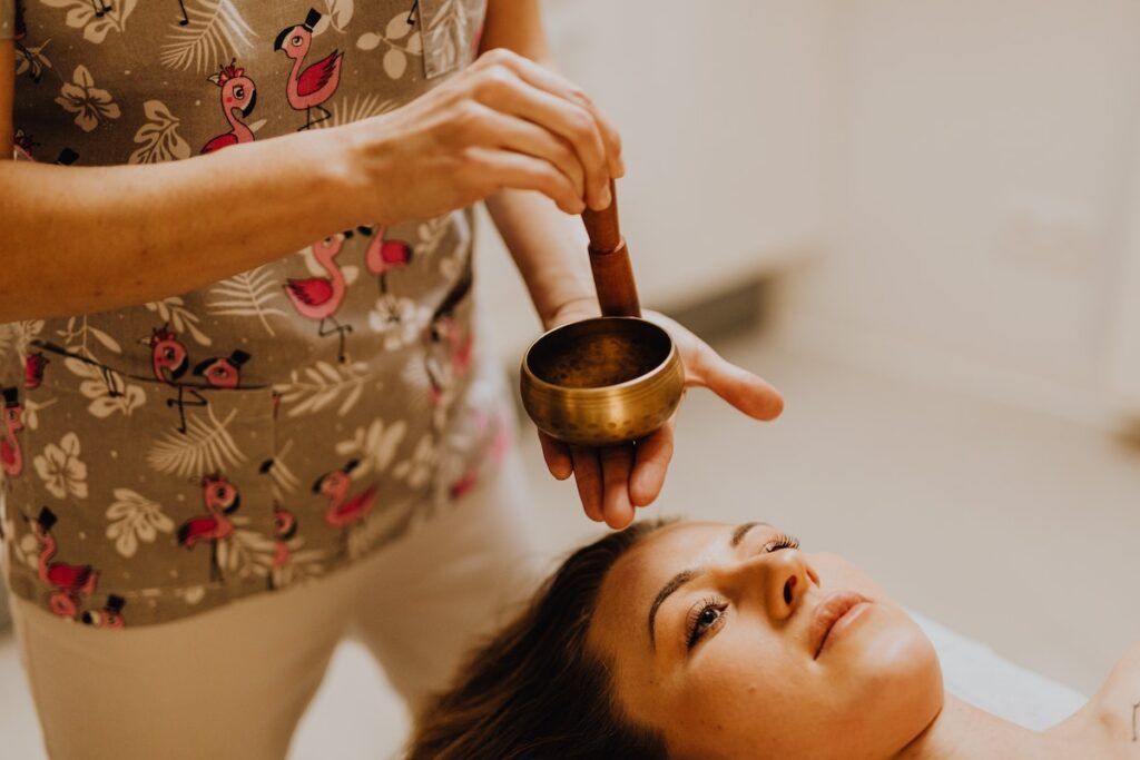 Woman lies in spa setting as technician uses sound healing bowls above her head