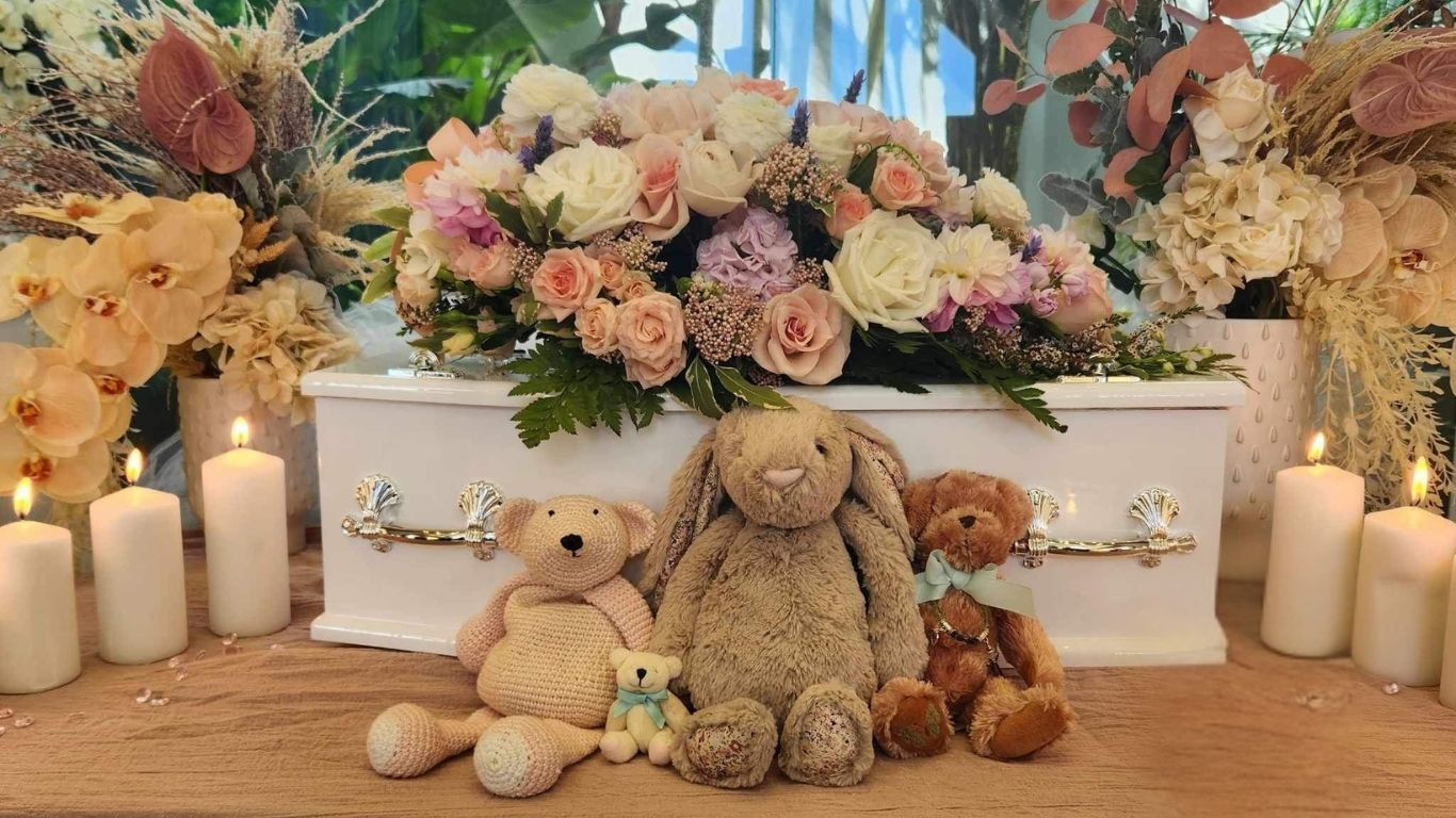 A white coffin with flowers and teddy bears on it.