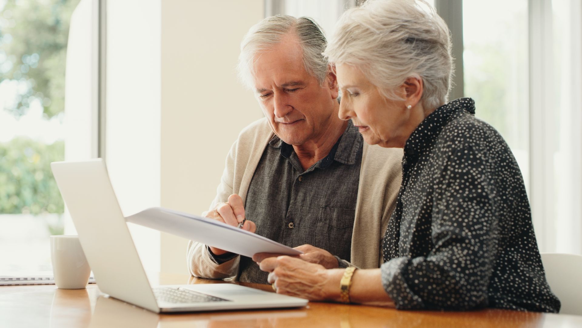 An elderly couple is sitting at a table looking at a laptop computer.