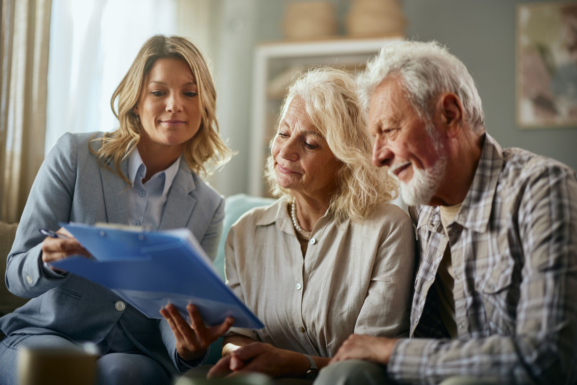 An elderly couple is sitting on a couch talking to a woman who is holding a clipboard.