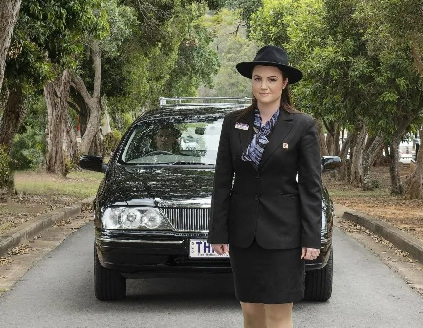 This is an image of a lady funeral director from Tony Hollands Funerals walking in front of a hearse in a cemetery
