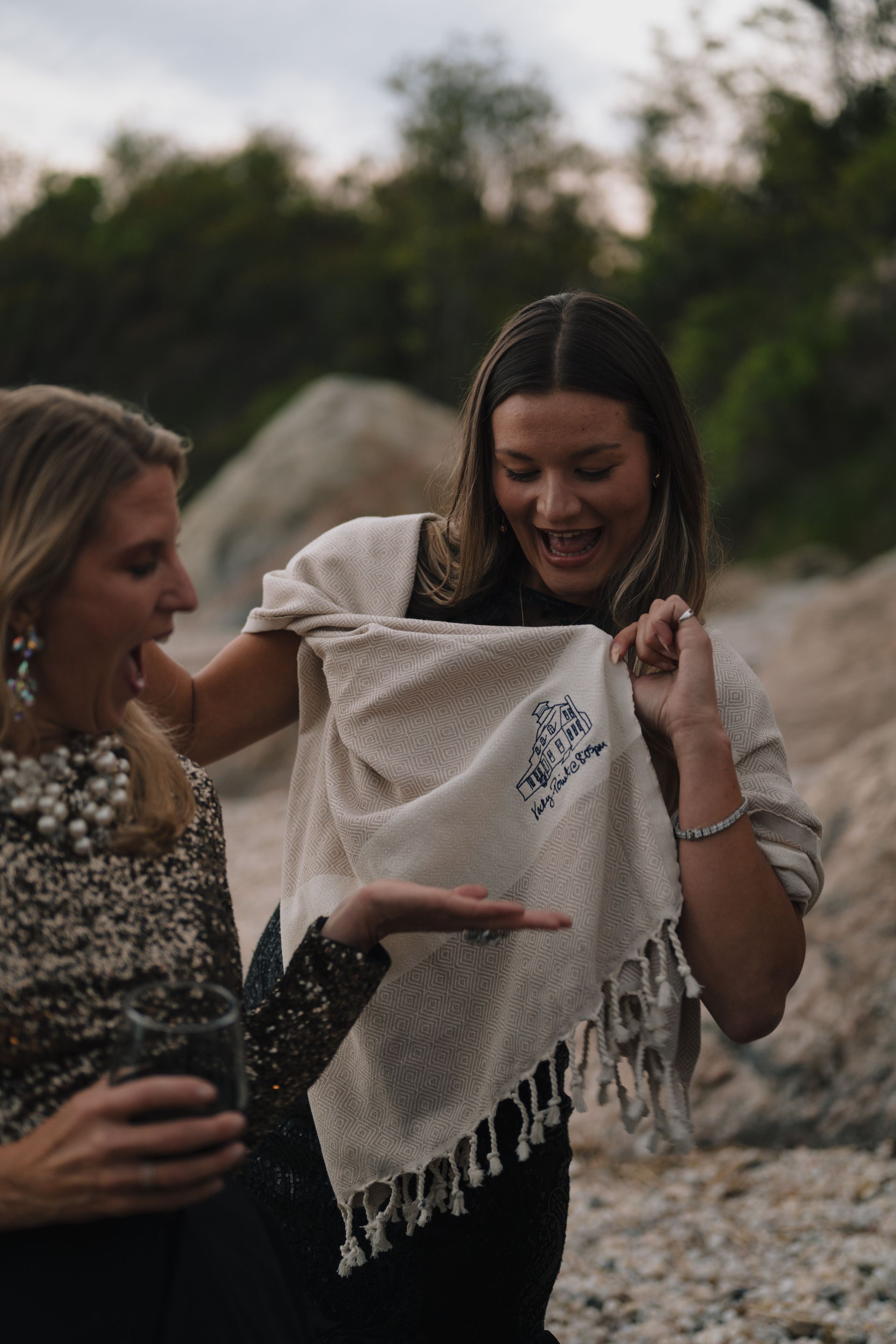 Two women outdoors examining a cream knit shawl with fringe on a rocky beach