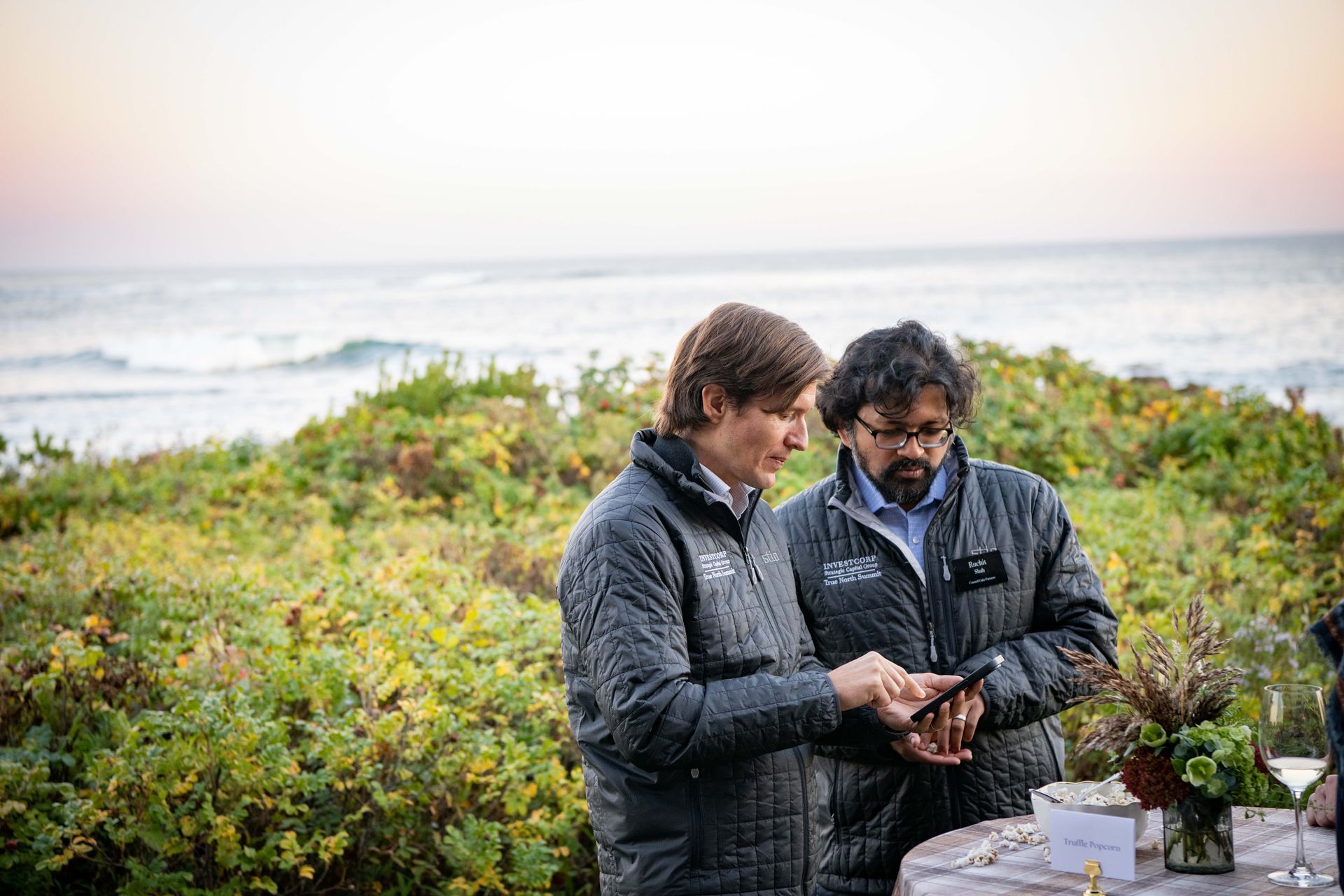 Two people examining plants in a coastal field, with ocean waves in the background.