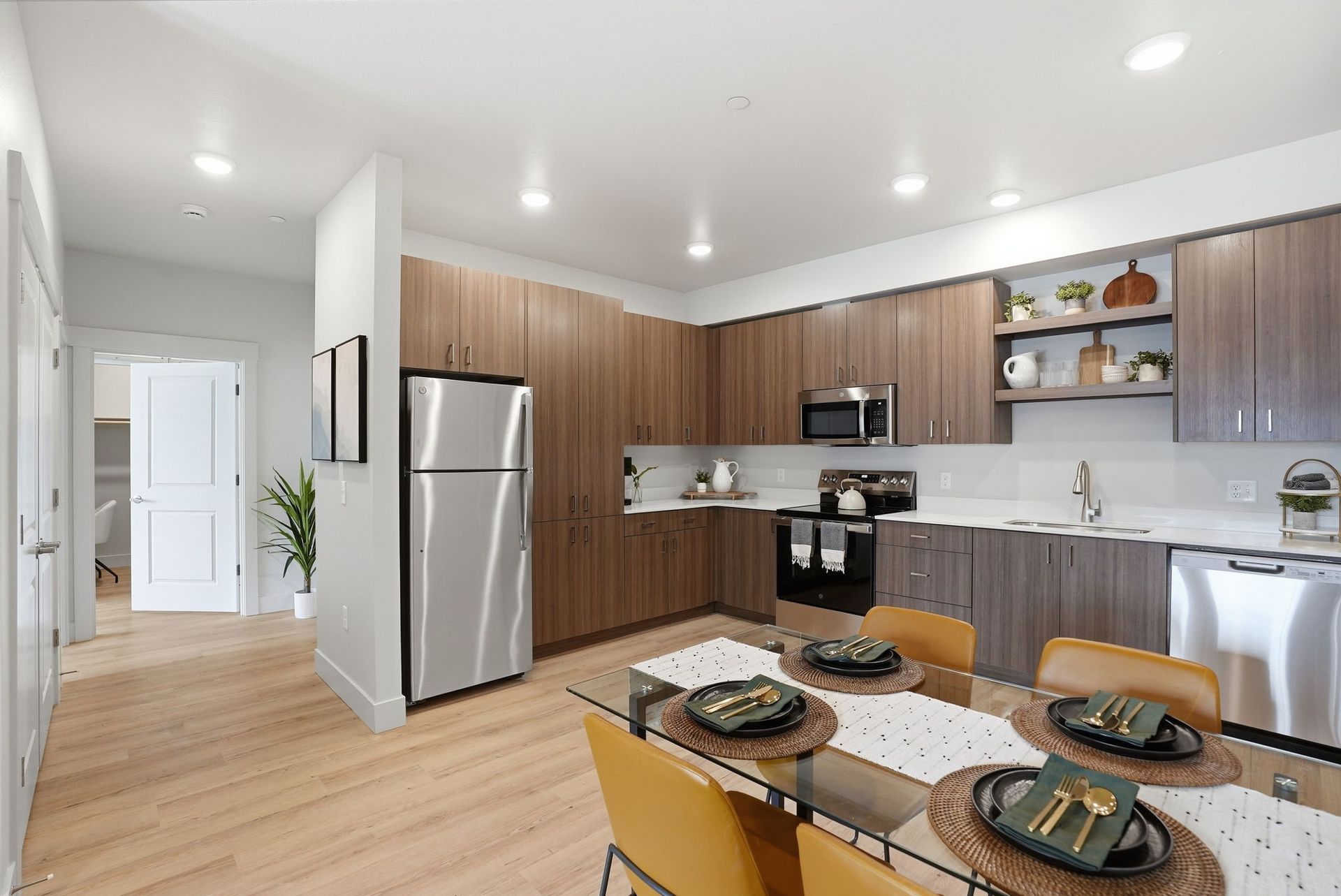A glass dining table and orange chairs sitting near a modern kitchen featuring wood cabinets and stainless steel appliances.