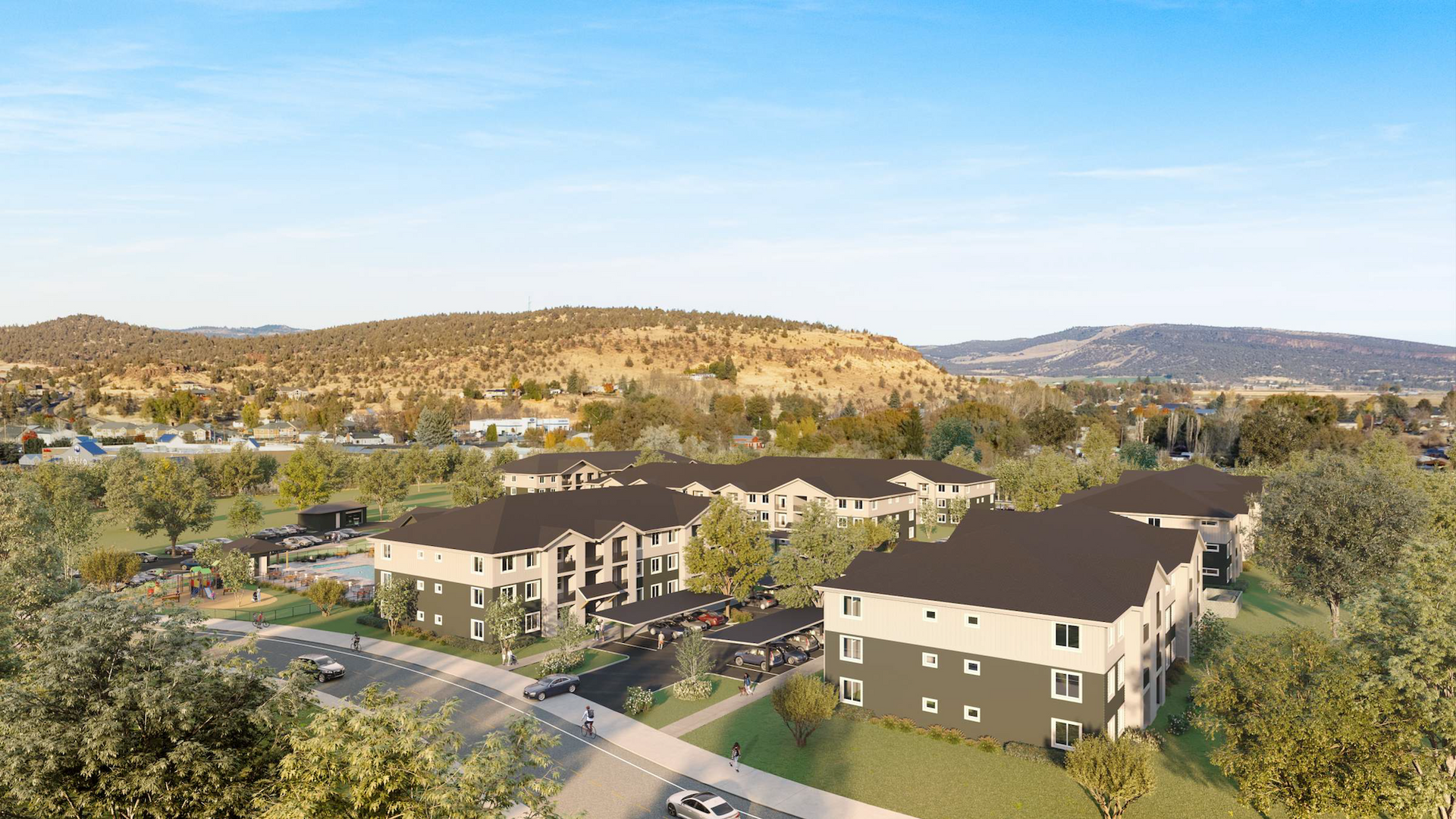 Aerial view of Mill at Ochoco apartment buildings with dark roofs nestled among trees on a hilly landscape.
