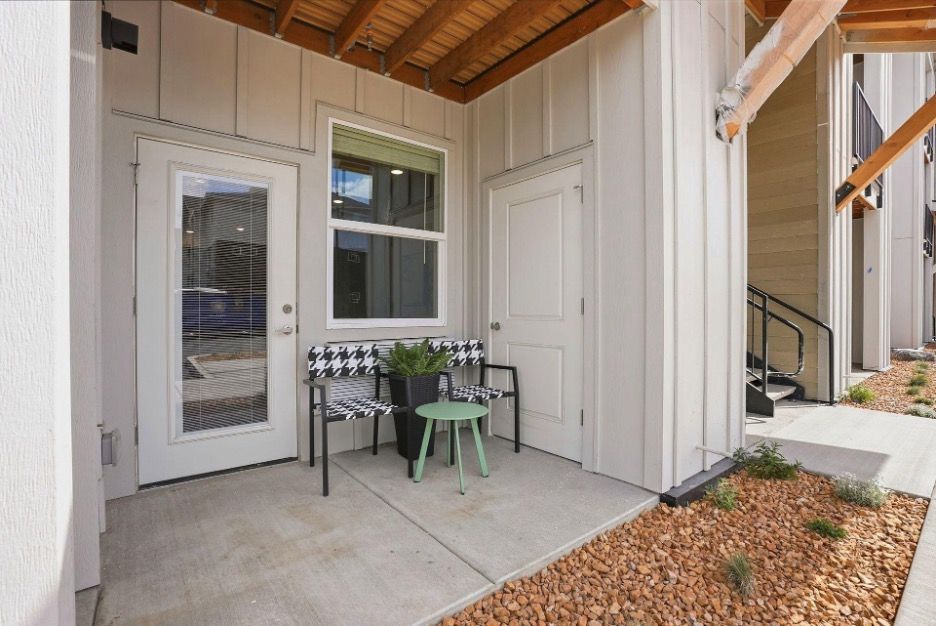 Covered front porch with two chairs, small table, and white siding beside a door and window.