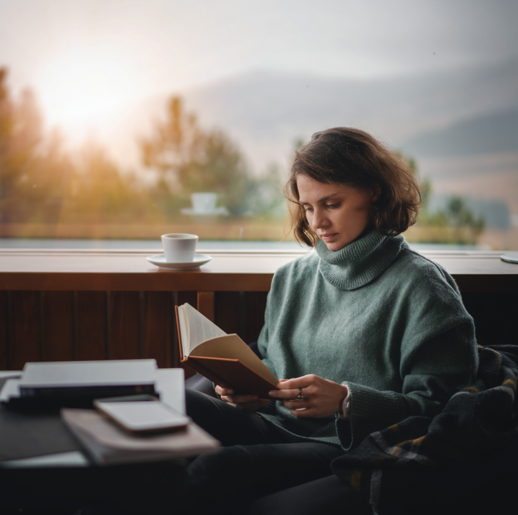 Woman in a green sweater reading a book by a sunlit window with a cup of coffee on the table beside her.