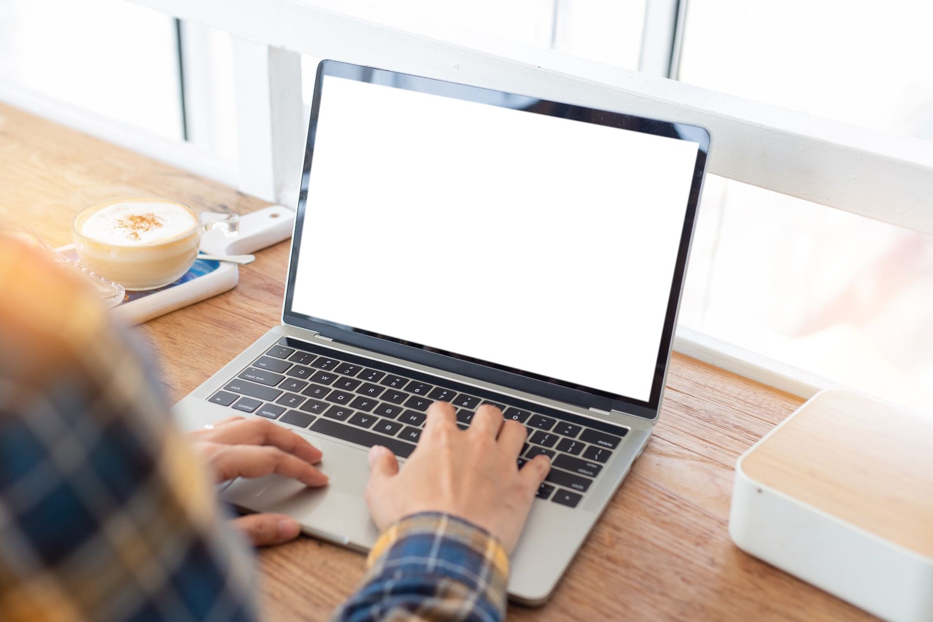 Person typing on a laptop with a blank white screen at a wooden desk near a bright window with coffee nearby.