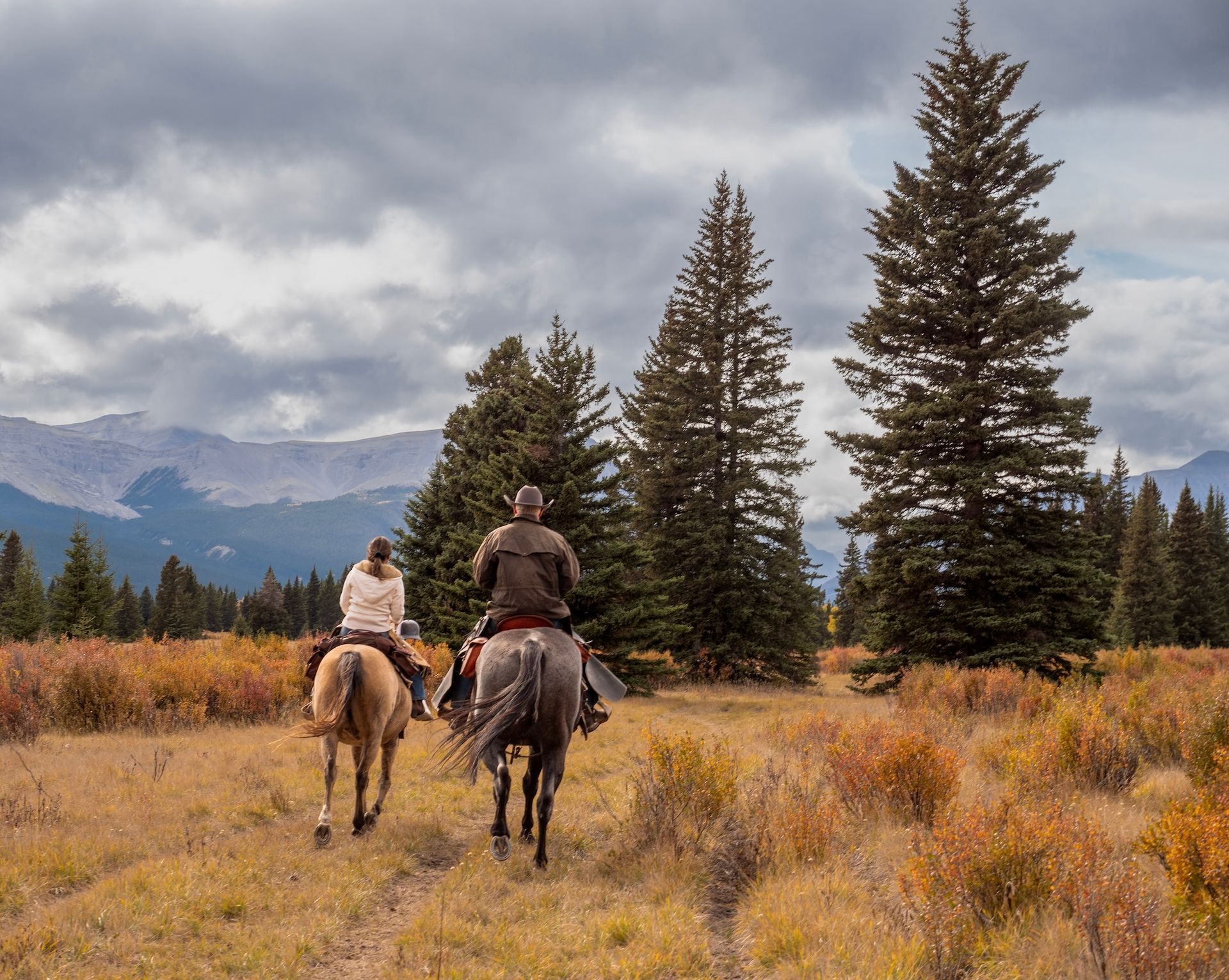 Two riders on horseback moving through a golden fall meadow with tall pines and mountain peaks behind them.