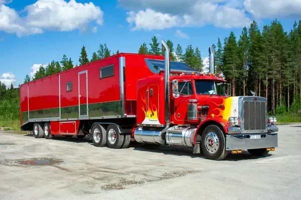 Red semi-truck with a large red trailer parked on a gravel lot near pine trees under a blue sky