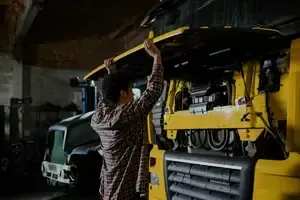 Person working inside a yellow truck engine bay in a dim garage, reaching up with a tool.