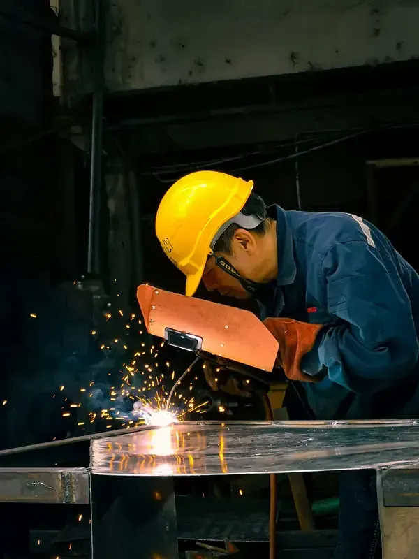Worker in yellow helmet and orange gloves welding metal, sparks flying in a dark workshop.