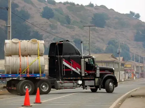 Black semi-truck carrying white tanks on a road beside orange cones and hills