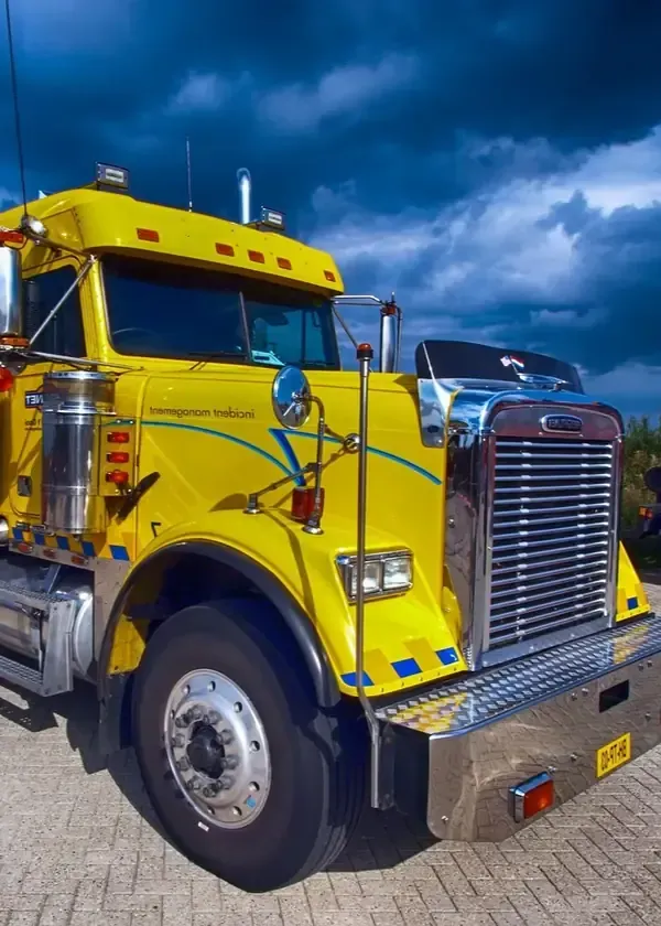 Bright yellow semi-truck with chrome grille parked on a paved lot under stormy blue clouds