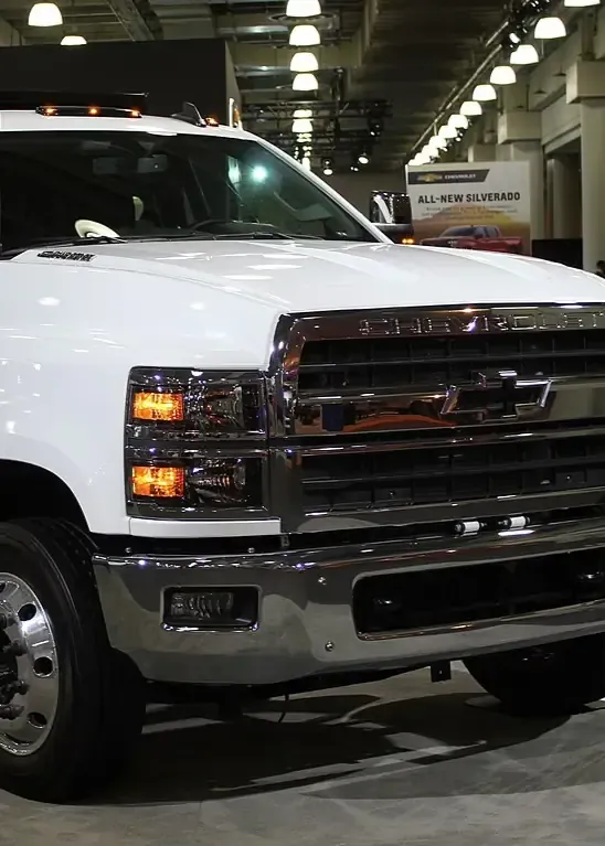 White pickup truck displayed indoors at an auto show, front view with headlights on
