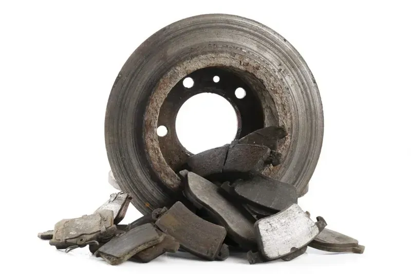 A worn metal car brake rotor surrounded by a pile of used, dark-colored brake pads against a plain white background.