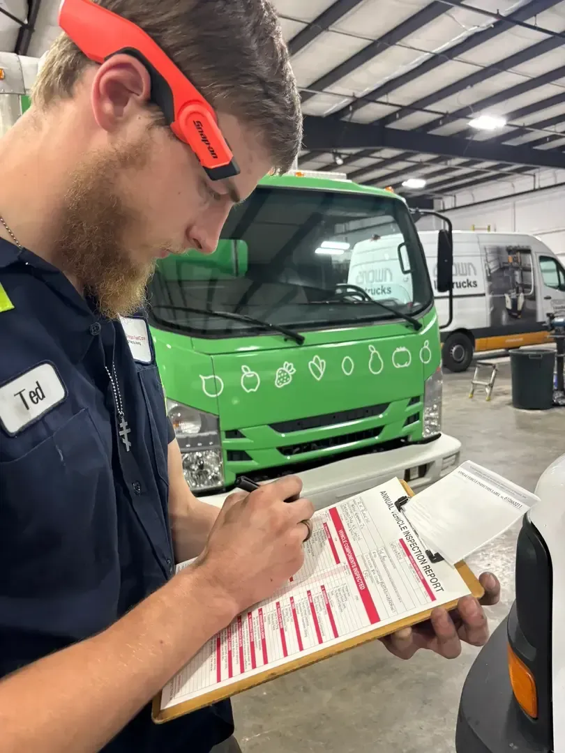Mechanic inspecting a silver car’s front fender with a clipboard in a garage