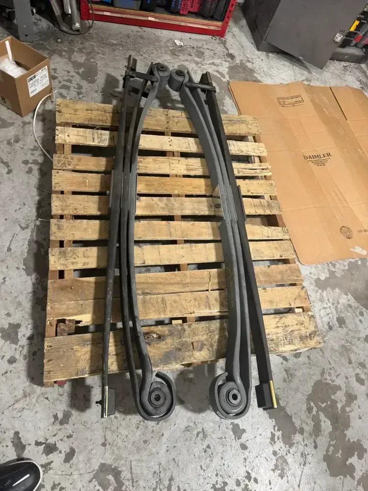 Black leaf springs stacked on a wooden pallet in a workshop floor area.