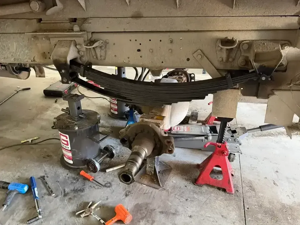 Vehicle axle and suspension under repair in a garage, with jack stands, tools, and a removed driveshaft on the floor.
