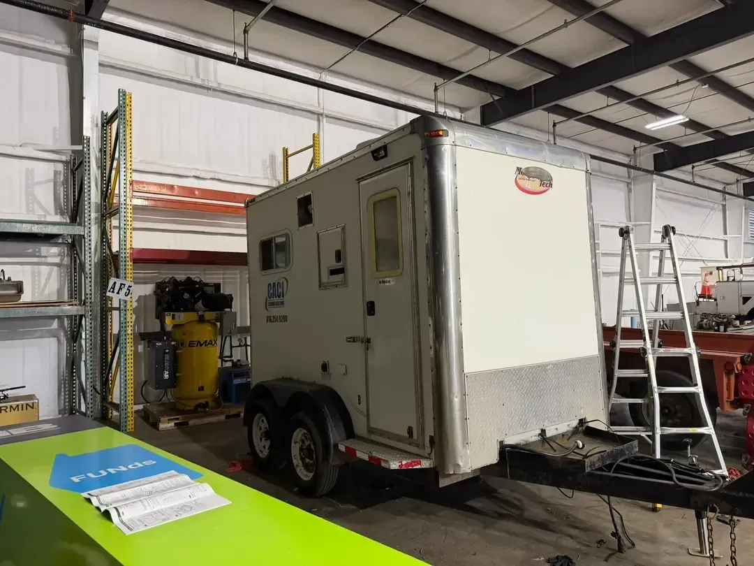 White utility trailer inside a workshop beside a ladder and shelving unit.