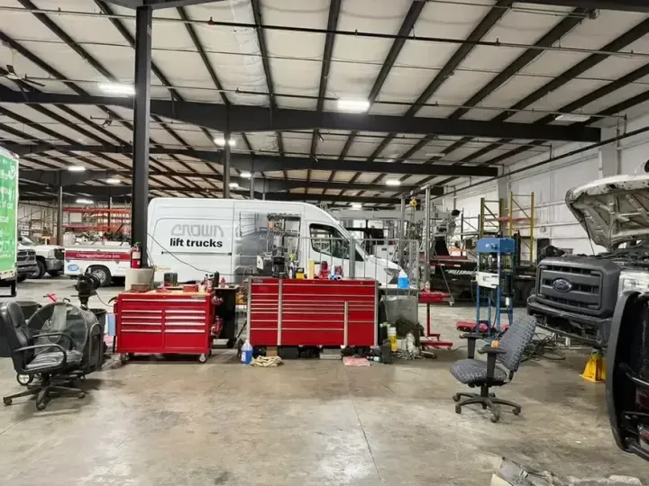 Person working on a large engine in a garage, with a white vehicle nearby.