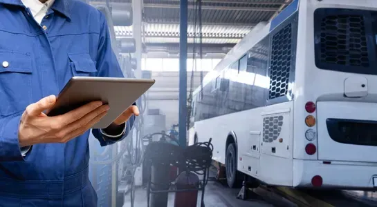 Technician in blue uniform using a tablet beside a bus in a maintenance garage