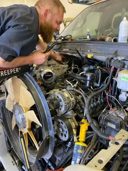 Mechanic leaning over an exposed car engine, inspecting wiring and components in a workshop.