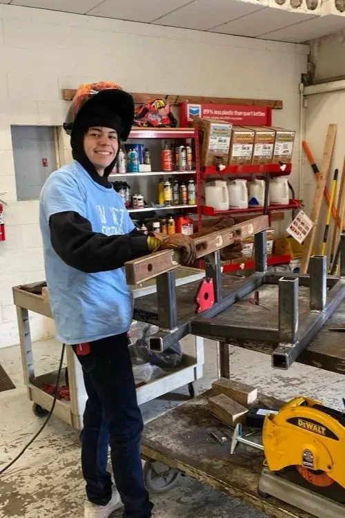 Person in a blue shirt and helmet stands in a workshop beside a metal frame and yellow saw.