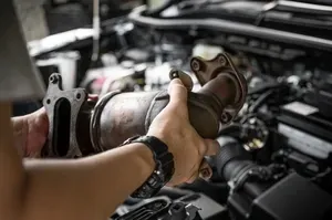Hands holding a car catalytic converter in an engine bay during repair