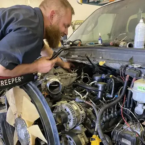 Mechanic in gray overalls inspects machinery under a vehicle's hood