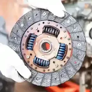 Gloved hands holding a circular car clutch disc with visible springs and textured friction surface
