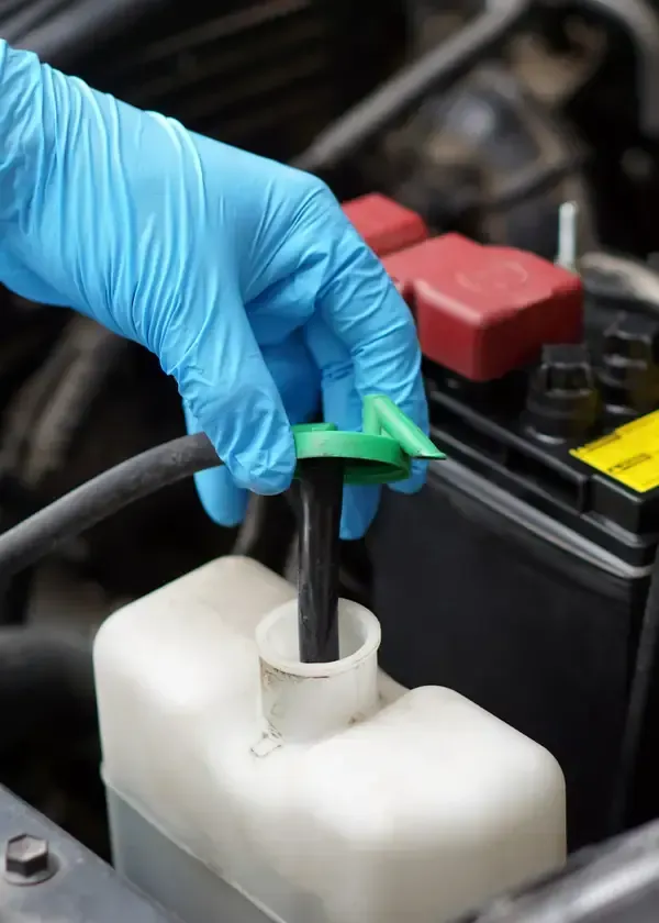Gloved hand inserting a hose into a white car fluid reservoir near a battery