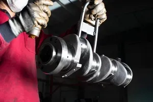 Worker holding a metal crankshaft in a dim workshop