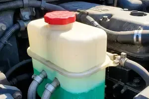 Car engine coolant reservoir with red cap and translucent white tank, partly filled with green fluid.