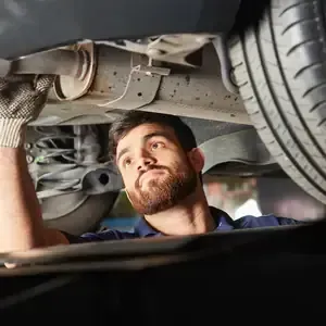 Mechanic inspecting a car’s undercarriage on a lift, looking upward with a focused expression