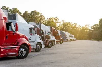 Line of parked semi trucks, including red and white cabs, beside a sunlit road at sunset