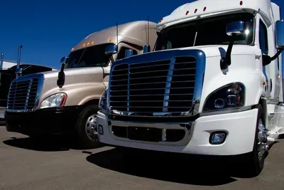 Two semi trucks, one white and one beige, parked side by side under a blue sky